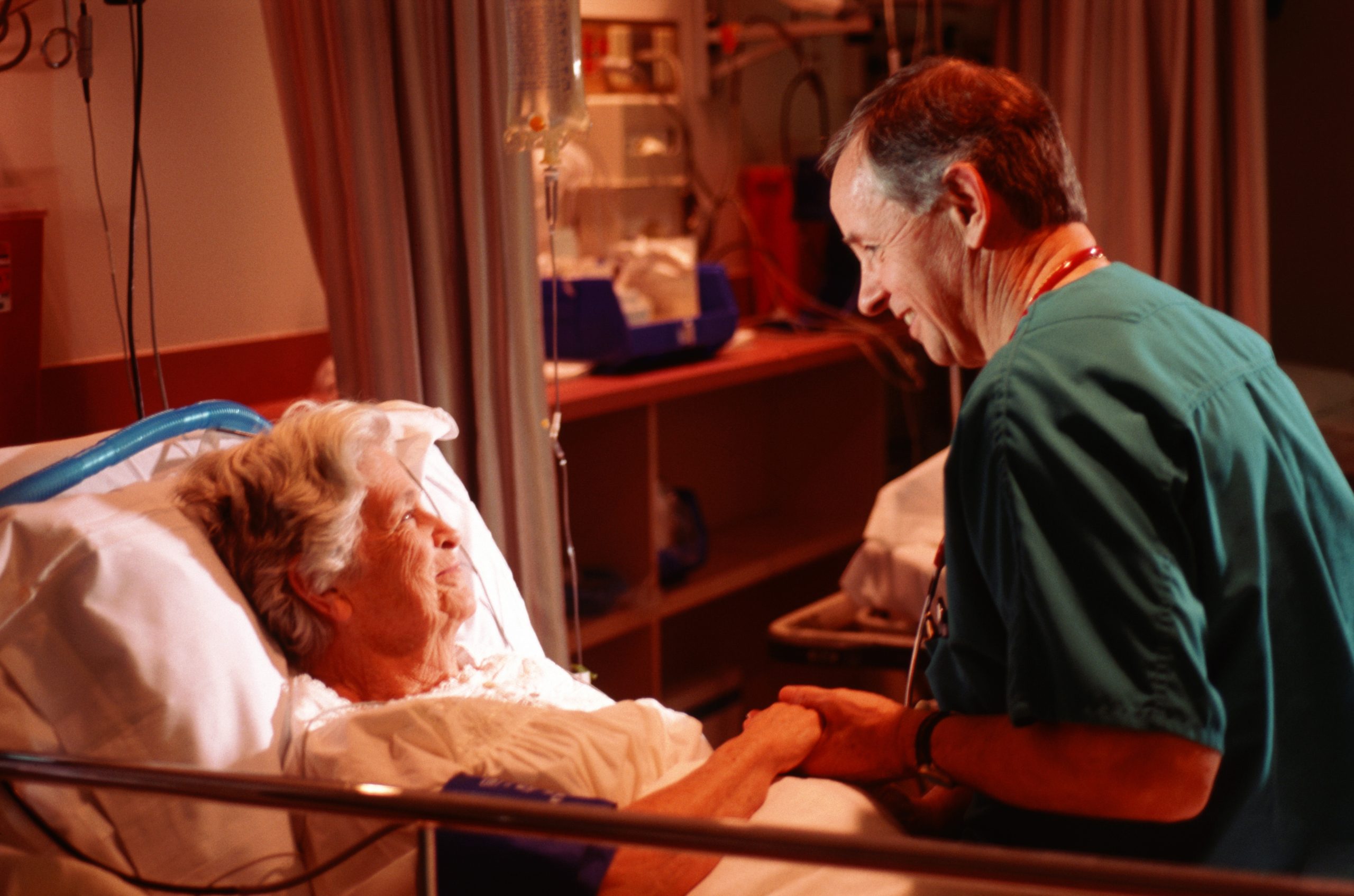 Ein Mann in medizinischer Kleidung hält die Hand einer älteren Frau im Krankenhausbett