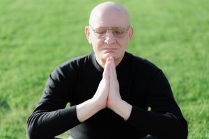Ein älterer Mann sitzt mit geschlossenen Augen und aneinandergelegten Händen im Grünen.
