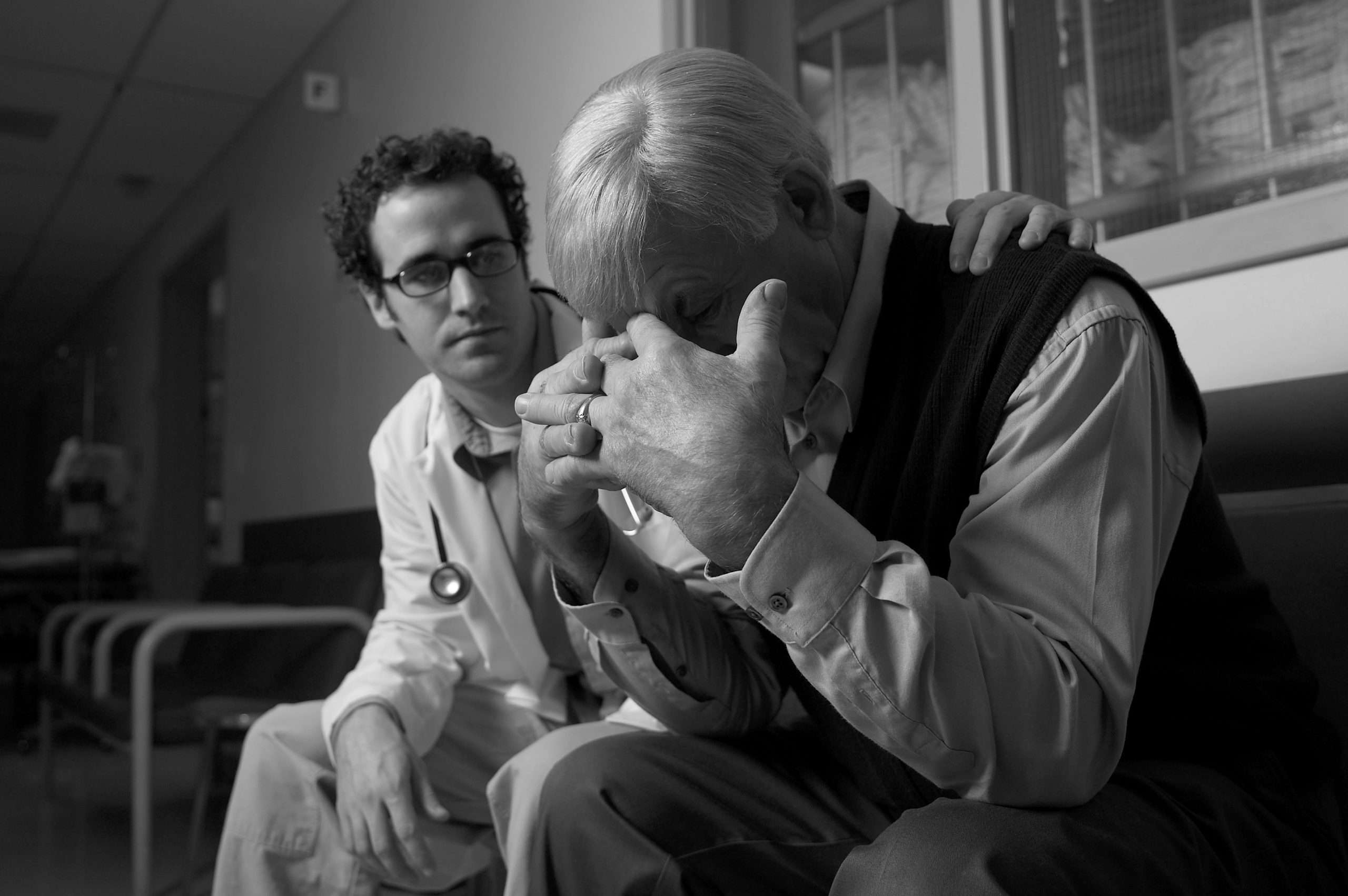 Schwarzweißfoto: Ein Mann mit Stethoskop legt einem älteren Mann behutsam die Hand auf die Schulter